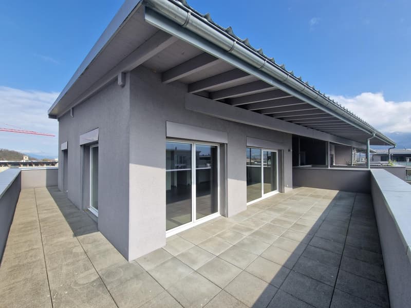 The image shows a modern apartment building with a large balcony or terrace. The building has a gray exterior with large windows and sliding glass doors leading out to the balcony area. The roof has a distinctive angled design with metal roofing. The balc