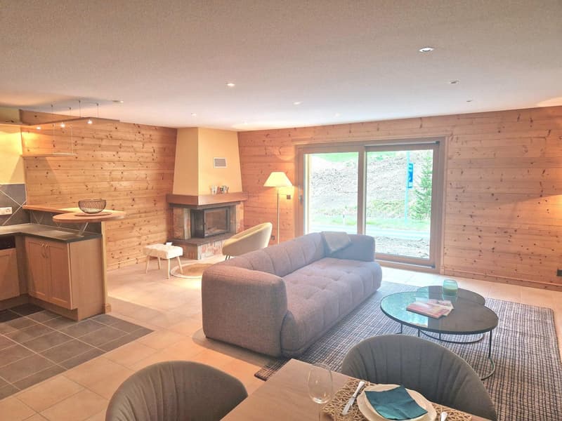 Spacious living room with wood-paneled walls, a fireplace, a large sofa, a coffee table, and a dining area with a table and chairs. The room has large windows overlooking the outdoor scenery.