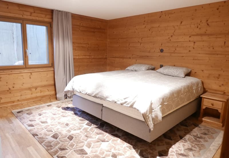 Spacious bedroom with wooden walls, large windows, a king-size bed, and a stone tile floor. The room has a cozy and rustic feel.