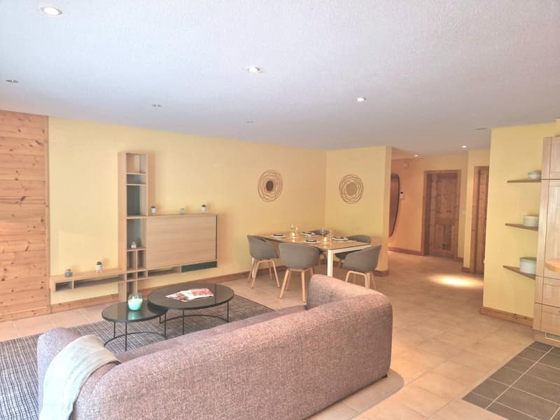 Open-concept living room and dining area with yellow walls, wooden accents, a gray sofa, a dining table with chairs, and a fireplace. The room also features shelving units, coffee tables, and decorative wall art.