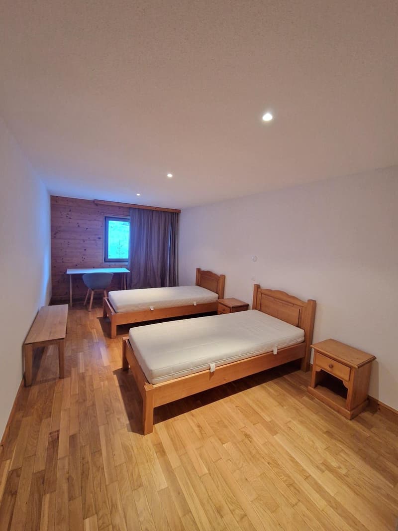 Spacious bedroom with wooden floor, two twin beds, wooden furniture including nightstands, and a window with curtains.