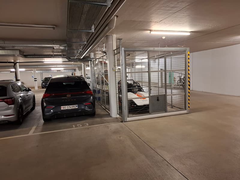 The image shows an indoor parking garage with concrete floors and walls. There are several cars parked in the garage, including a black car in the foreground. The garage has a wire mesh enclosure, likely for security purposes.