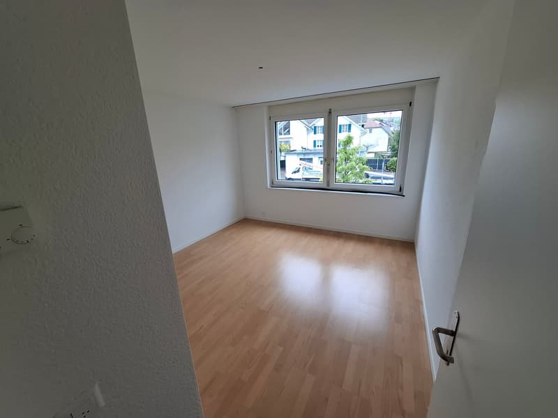 Spacious bedroom with hardwood flooring, white walls, and a large window providing natural light.