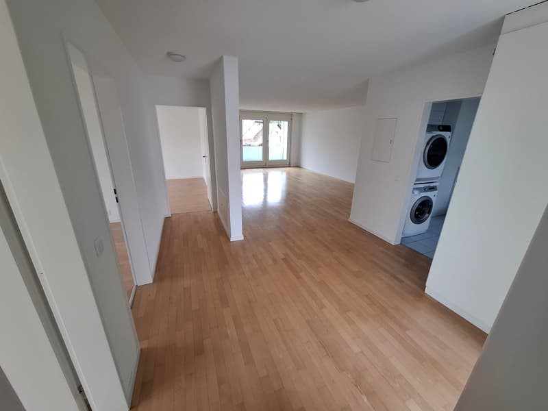 Spacious open-plan living area with hardwood floors, white walls, and large windows. The apartment includes a laundry area with a washing machine and a dishwasher.
