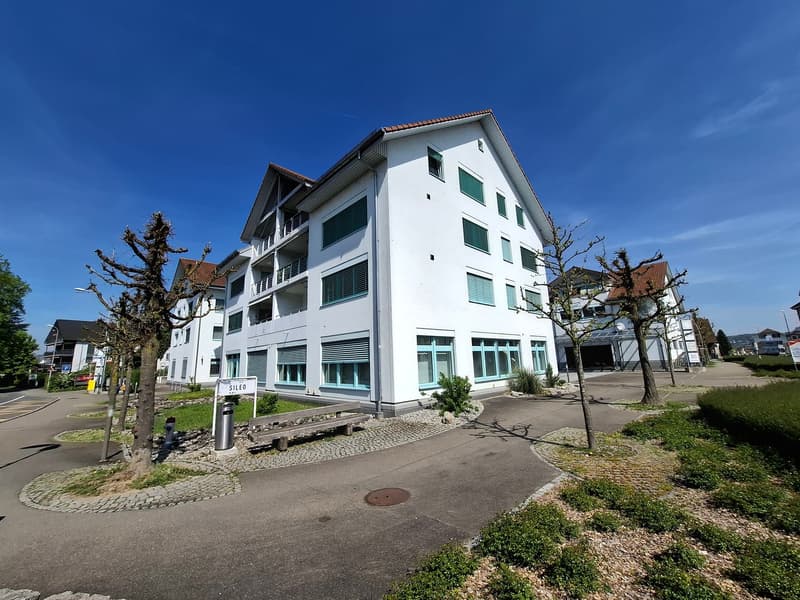 A multi-story white apartment building with balconies, surrounded by trees and a paved walkway. The building has a modern, minimalist design with large windows.