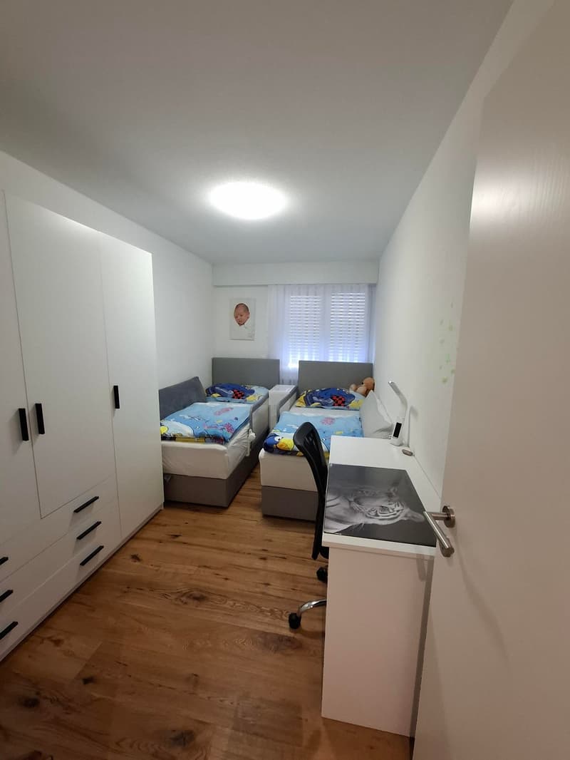 Children's room with two beds, desk, and wardrobe. White walls and wooden floors.