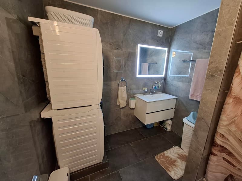 modern bathroom with tiled floor, vanity unit with sink, mirror, and shelves, drying rack, built-in shower, toilet, and washing machine
