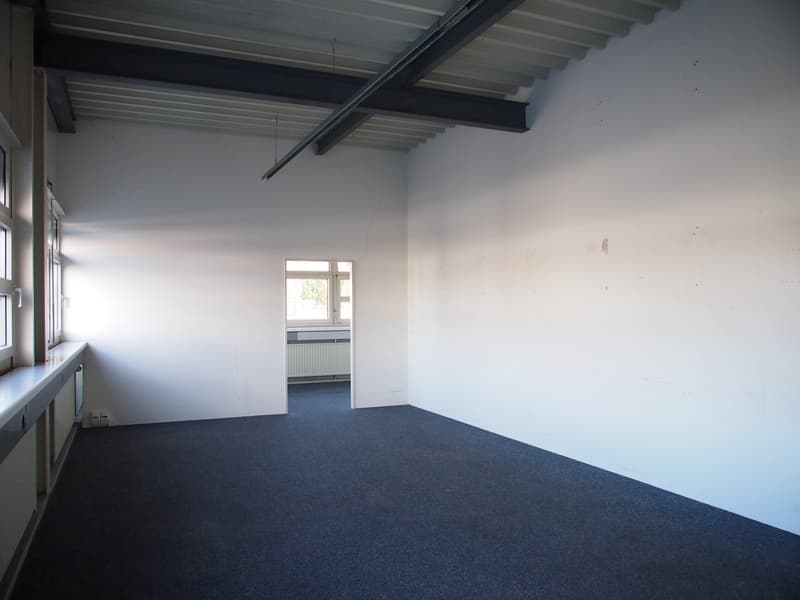 Empty room with white walls, exposed ceiling beams, and a dark blue carpeted floor. The room has a window and appears to be a commercial or office space.