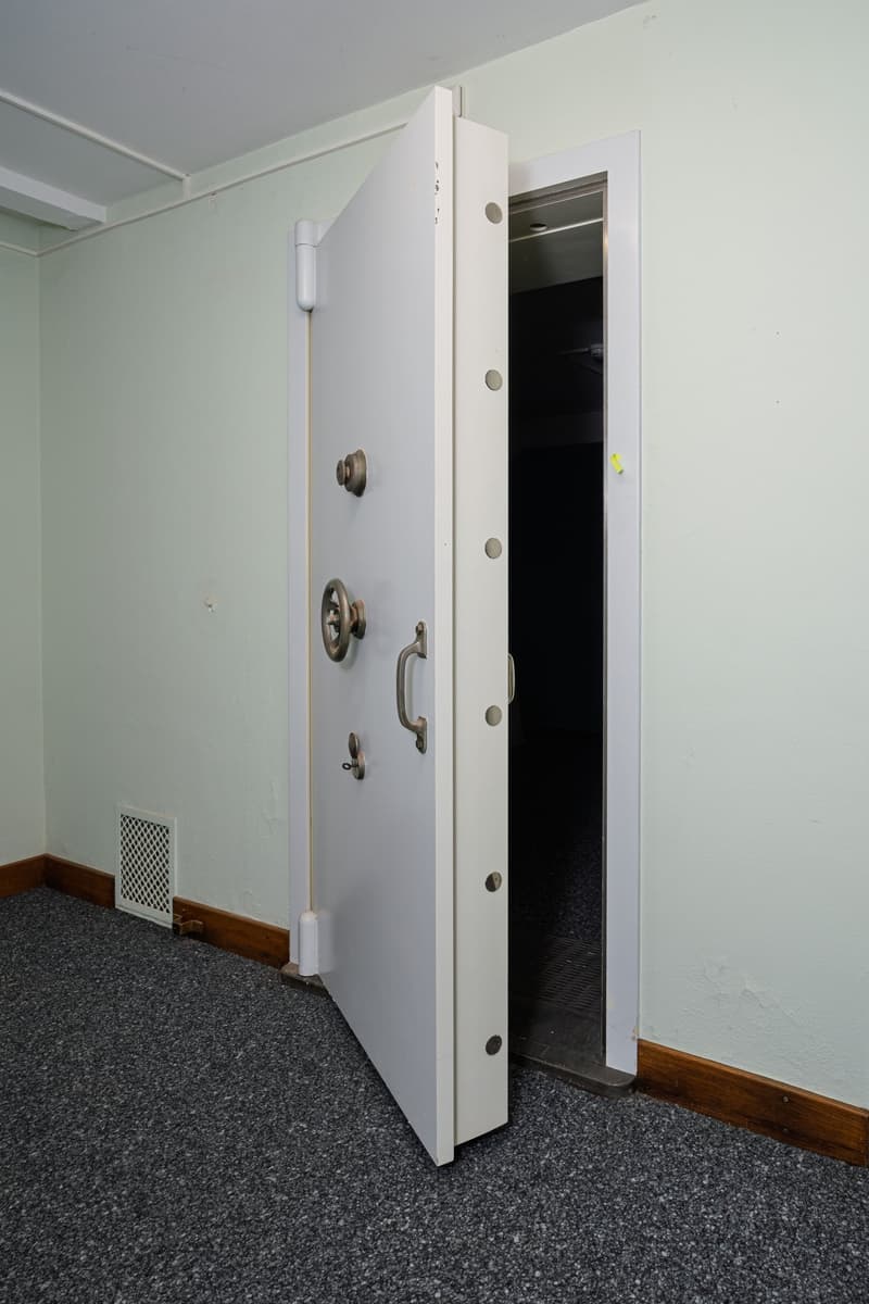 The image shows a white metal security door or safe in a room with a gray carpeted floor. The door has various locks and handles, indicating it is a secure entry point.