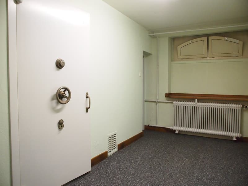 The image shows a room with white walls, a radiator, and a wooden cabinet or shelving unit. The floor appears to be covered in a dark-colored, textured flooring material, possibly a type of linoleum or vinyl. The room has a simple, utilitarian appearance.