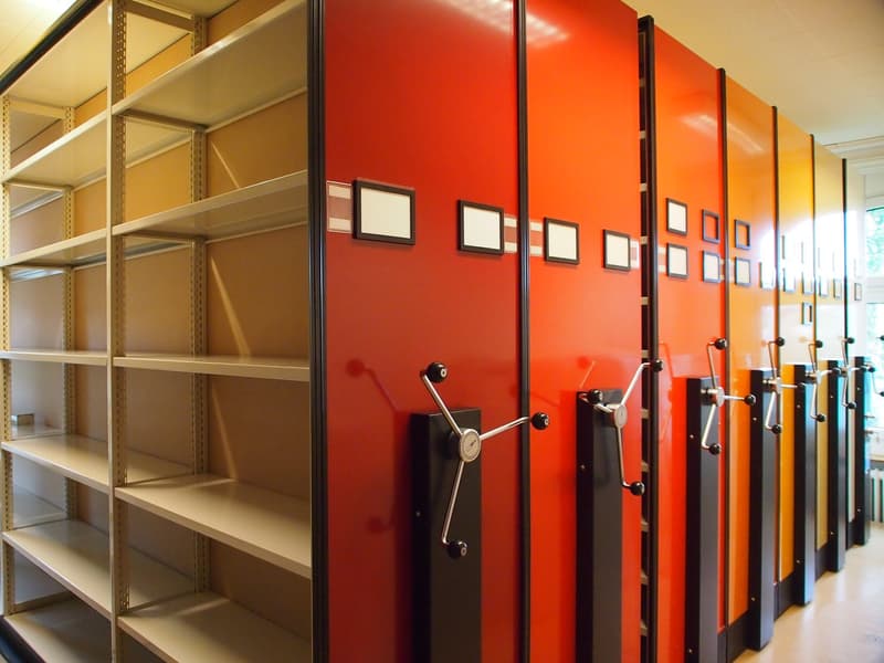 The image shows a row of bright orange storage lockers with metal shelves on the left side. The lockers have locks and handles, and there are framed pictures hanging on the wall above the lockers.