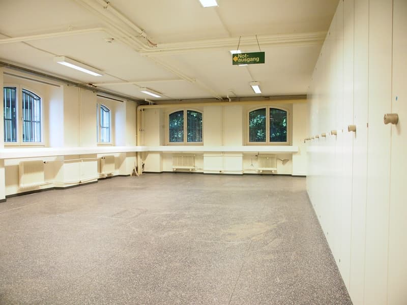 Large open room with high ceilings, arched windows, and a 'Not ausgang' (emergency exit) sign. The room has a concrete floor and appears to be a multipurpose space, possibly used for events or activities.