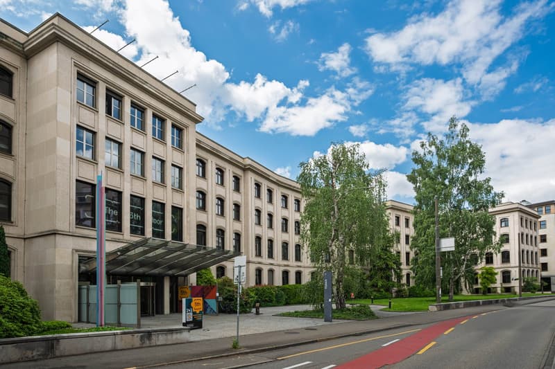 Large multi-story building with a classical architectural style, featuring rows of windows and a covered entrance. The building is surrounded by trees and greenery, with a paved area in front that appears to be a parking lot or plaza. The sky is blue with