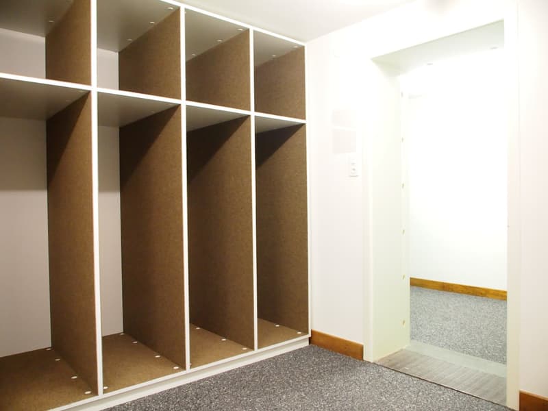 The image shows a bedroom with a built-in wardrobe or closet system. The wardrobe has multiple compartments or cubbies made of a brown material, likely wood or a wood-like material. The room has white walls and a gray carpeted floor.