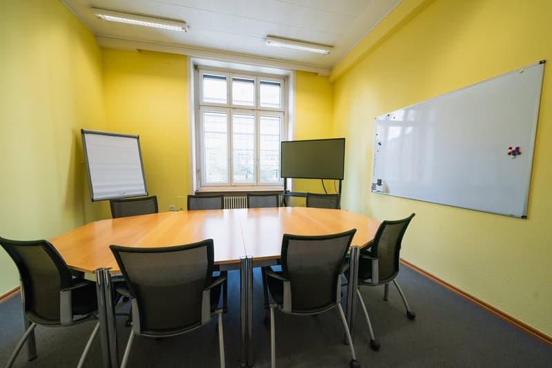 A conference room with yellow walls, a round table, black chairs, whiteboards, a monitor, and a radiator.