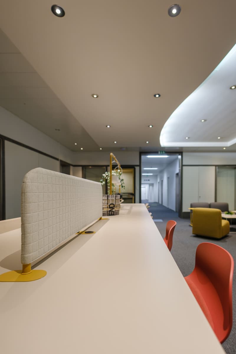 Modern office layout with long white desks, gray walls, red and yellow chairs, and ceiling lighting