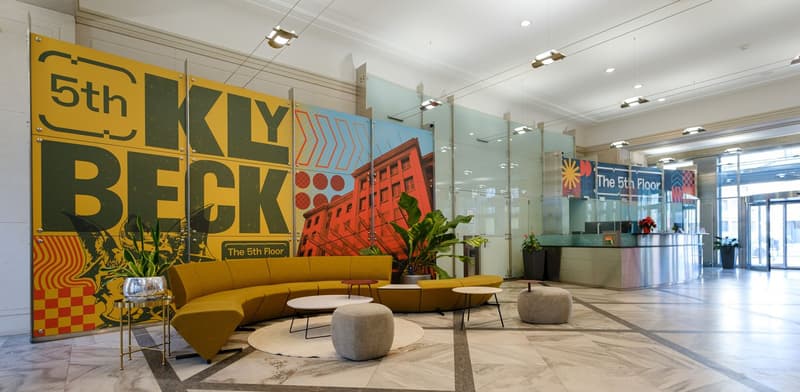 A modern lobby with a yellow sofa, coffee tables, circular ottomans, potted plants, and a large wall mural.