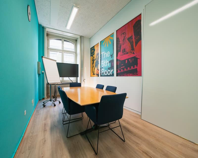 A clean meeting room with wooden table, blue chairs, whiteboard, monitor, and three paintings on the wall