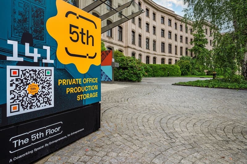 brick building with many windows, cobblestone ground, greenery and trees, sign that reads 'The 5th Floor' and 'PRIVATE OFFICE PRODUCTION STORAGE', QR code on the sign, large open area in front of the building