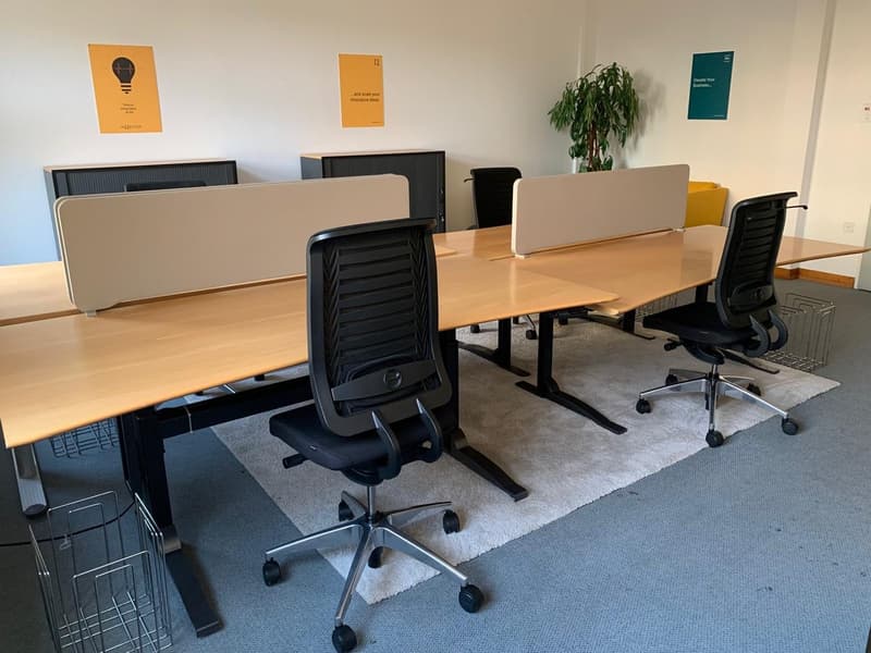 Empty office space with desks, office chairs, wall-mounted frames, and a potted plant.