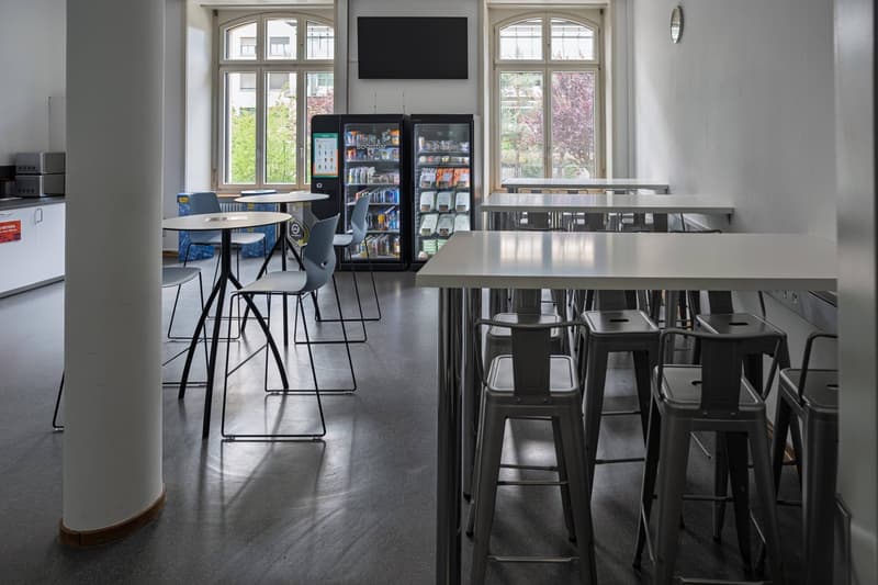 An empty room with tables and chairs, a large windows on the wall, two vending machines, and a television