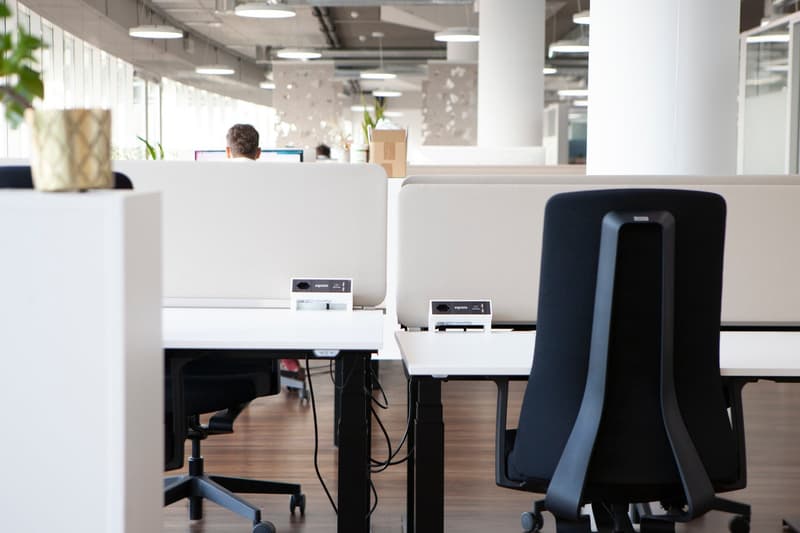 Modern office space with desks, chairs, monitors, plants, lamps, and people