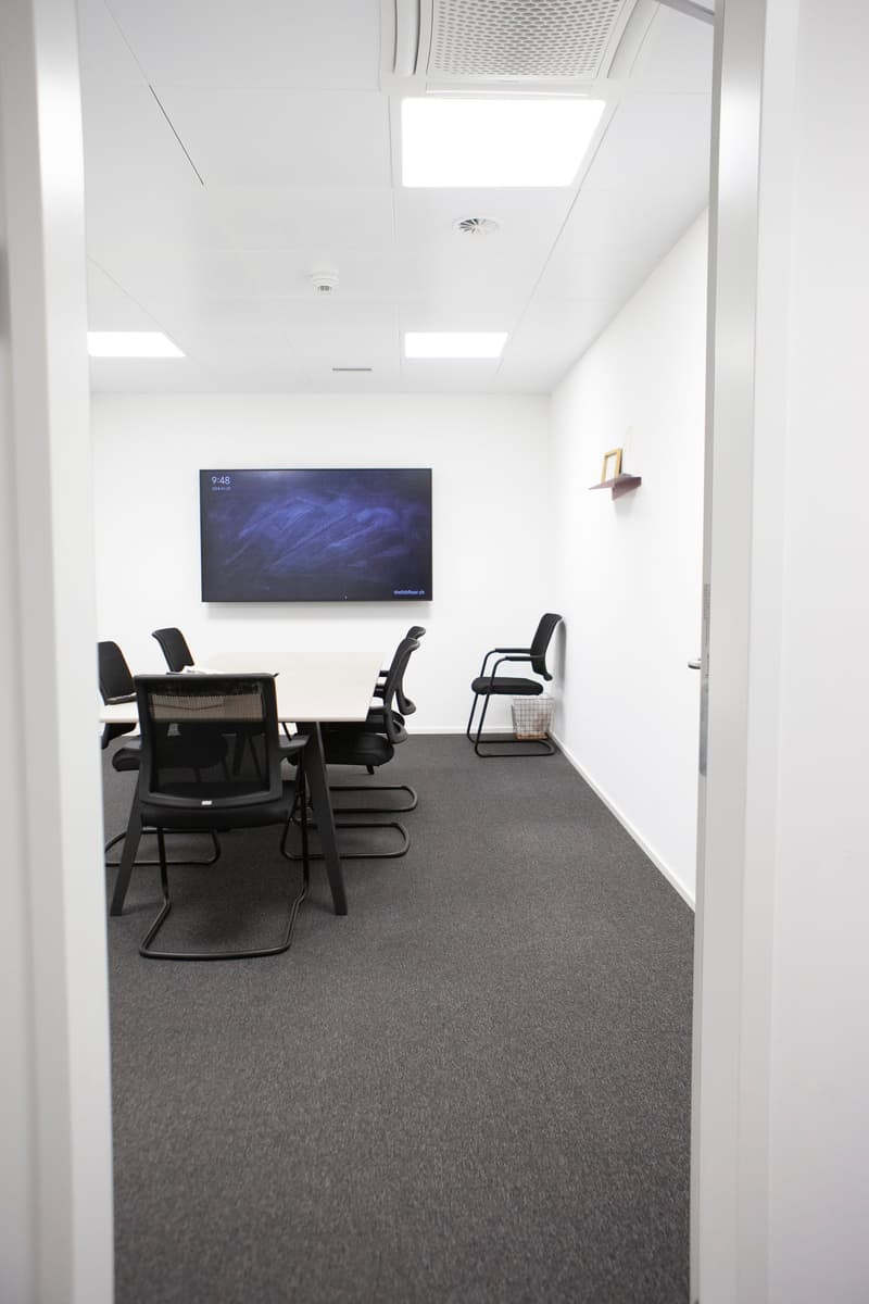 white walls, gray flooring, large meeting table, black chairs, flat screen mounted on the wall, small shelf on the wall