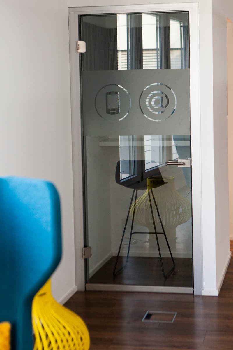 The image shows a room with a glass door, an empty office chair, and some reflective furniture. The door is closed, has a white handle, and some text and symbols are etched on it. The floor is made of wood. A blue and yellow chair is placed in front of th