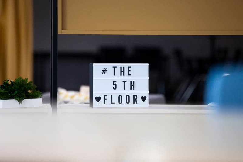 Desk with a lightbox that reads 'THE 5TH FLOOR' in front of a yellow curtain, potted plants on the desk.