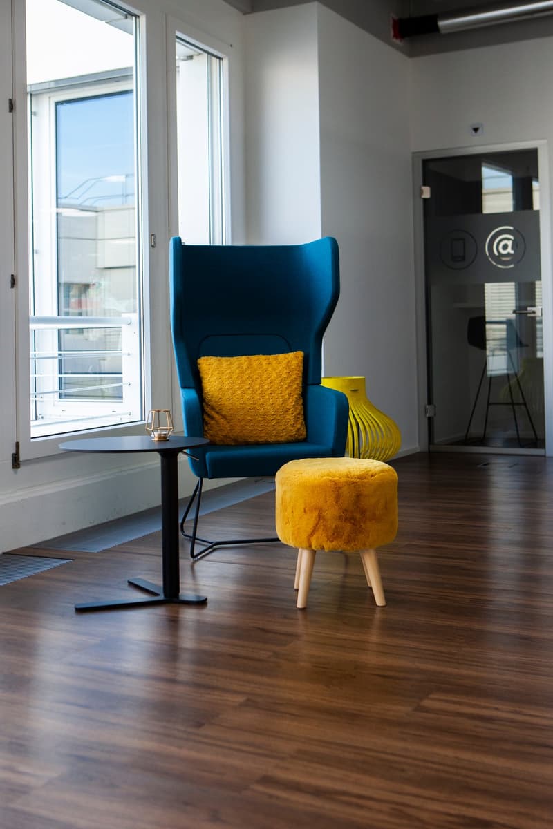 room with wooden floor, blue chair with yellow pillow, round yellow ottoman, small round table, glass window with open balcony
