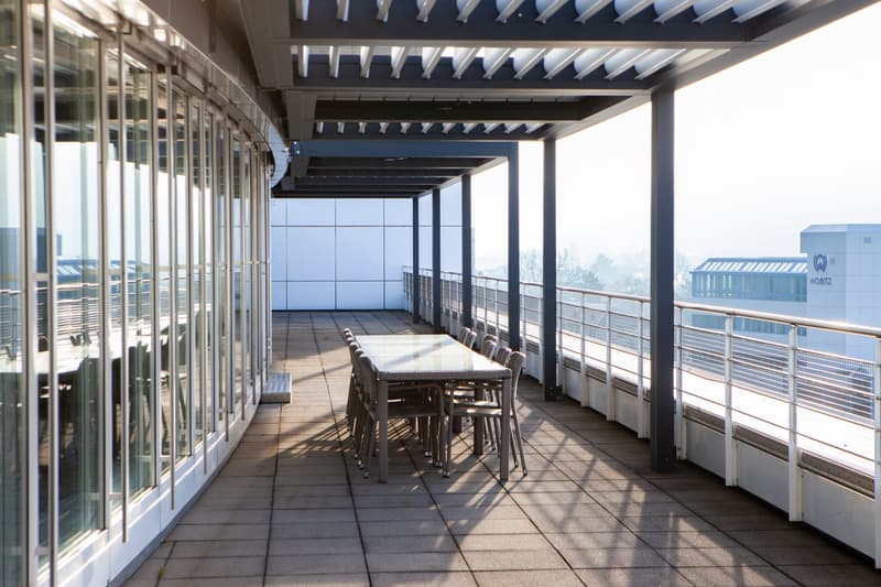 An open balcony/terrace with a large glass wall, tables, chairs, metal railings, and a building view.