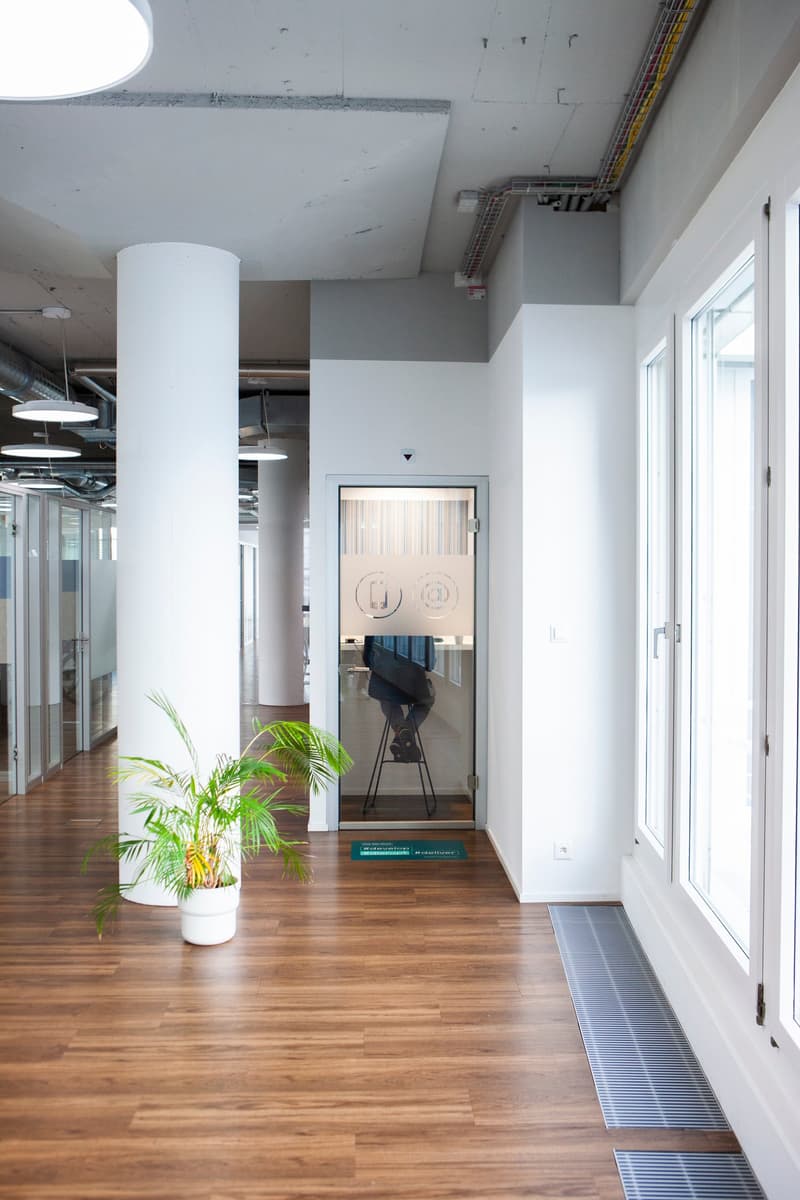 modern office hallway with wooden flooring, white walls, glass doors, ceiling lights, large column, and potted plant.