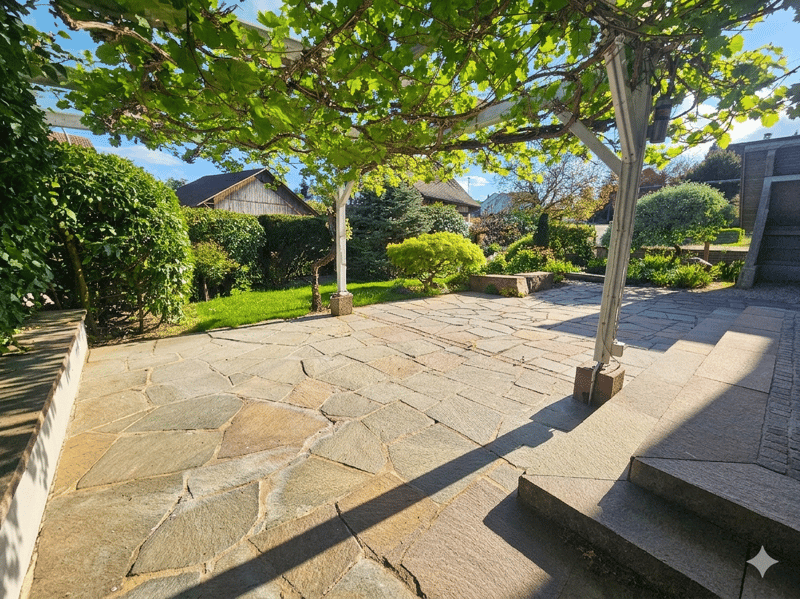 Stone paved patio with pergola, surrounded by shrubs and plants, steps leading to the patio, and a grassy area in the background.