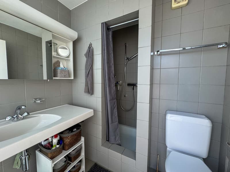 Gray tiled walls, white sink, mirror above sink, towel hanging on wall, white toilet, shower with silver showerhead