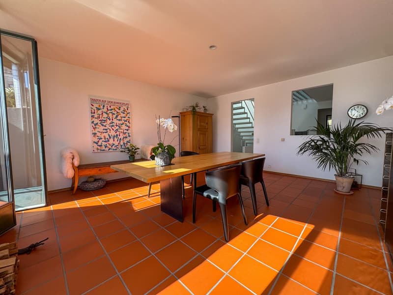 Dining area with orange tiled floor, large wooden table, black chairs, potted plants, artwork on wall, bench, clock, cabinet, staircase