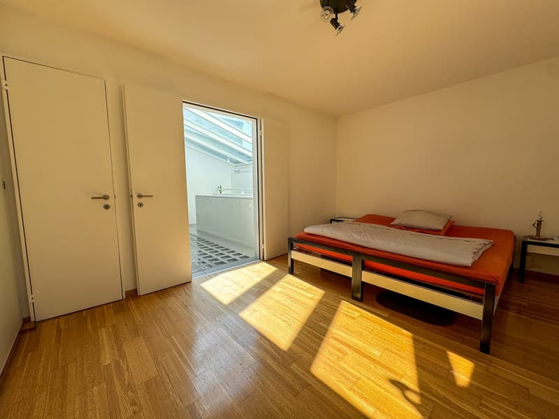 A bedroom with a bed, white doors, and a skylight window leading to the bathroom. Wooden floor, two white walls, and one wall with light shining through.