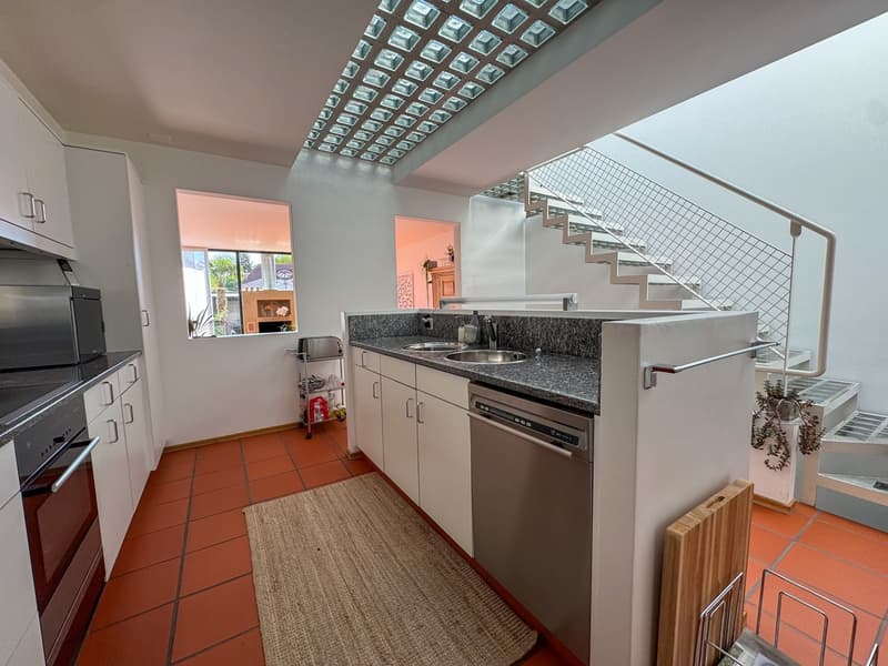 Modern kitchen with white cabinets, granite countertops, stainless steel sink, dishwasher, oven, and tiled floor. A staircase and window with natural light.