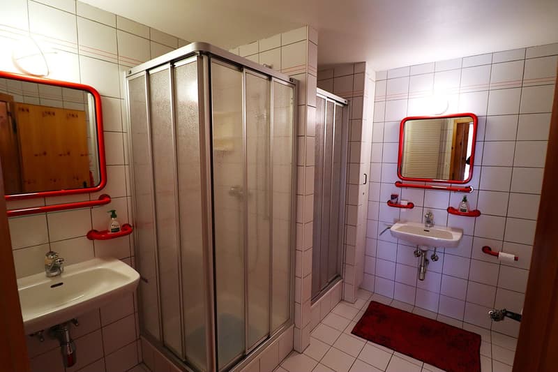 White tiled walls, shower with door, sink, mirrors with red frames, small red rug, towels