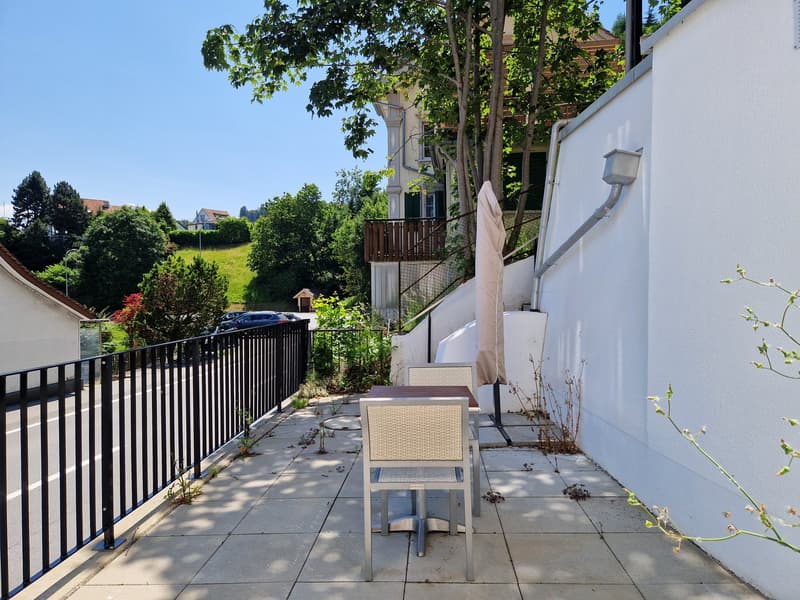 Outdoor dining area with table and chairs, tiled floor, black railing, trees, and plants