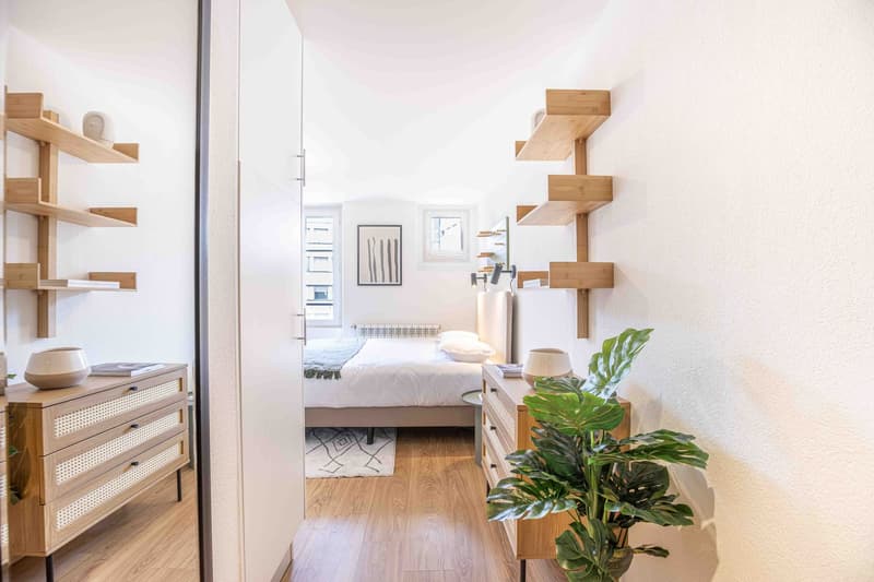 Spacious bedroom with white walls, wooden shelves, wooden dresser, and a large window. The room features a bed, a potted plant, and a framed artwork on the wall.