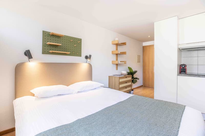 The image shows a modern and minimalist bedroom. The room has white walls and a large bed with a gray and white striped comforter. There is a wooden shelving unit on the wall with decorative items, and a wooden dresser with drawers next to the bed. The ro