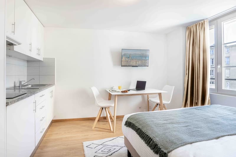 Bright and modern studio apartment with a kitchenette, a dining table, and a bed. The room has large windows, hardwood floors, and a TV mounted on the wall. The apartment appears to be designed for efficient living with a compact layout.