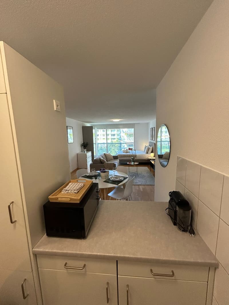 Compact kitchen with white cabinets, granite countertop, small dining area with round table and chairs, microwave and toaster on counter, kitchen island, mirror, picture frame, open living room with sofa, coffee table, bed, side tables, windows with view