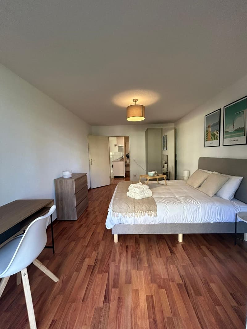 bedroom with a double bed, wooden floor, a desk, a chair, a night stand, lamps, a cabinet, a wall decor, and a kitchen view