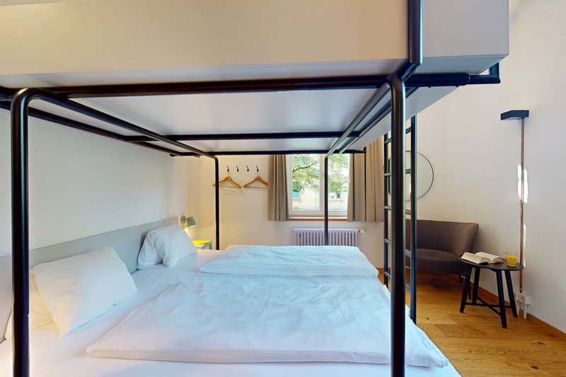 Double bunk beds, white sheets and pillows, nightstand with lamp, small couch, radiator, round mirror, power outlet, natural light from window.