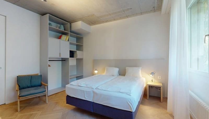 Wooden floor, white walls, white bed with pillows, wooden side table, lamp, wooden chair, closet with shelves, radiator, windows with white curtains