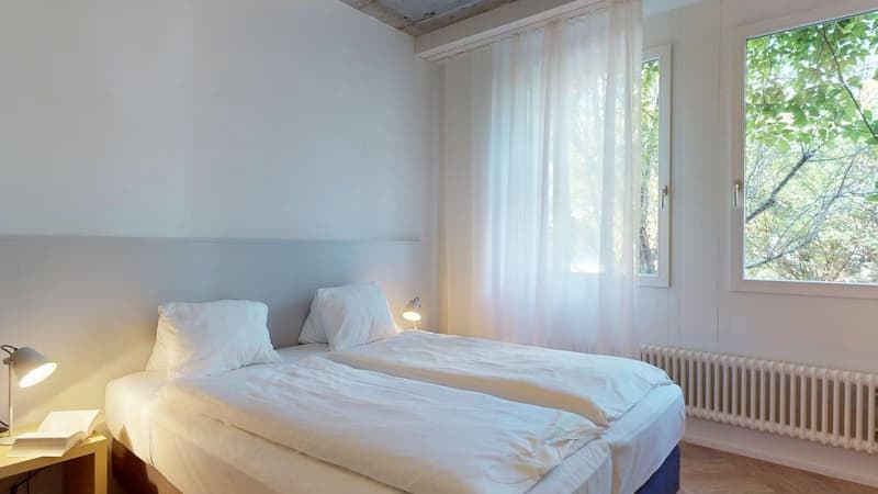 Two single beds with white bedding, a desk with a book, table lamps, curtains, wooden floor, and a window with a view