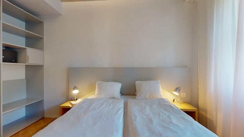 a bed with two pillows, lamps on the side tables, wooden floor, and wall with a shelf.