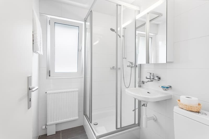 Bright and modern bathroom with a glass shower enclosure, white tiles, a sink with a vanity, and a window providing natural light.