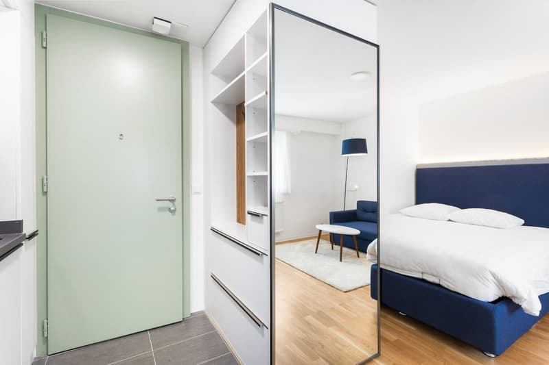 The image shows a modern and minimalist bedroom with a large mirrored wardrobe, a blue upholstered bed, a small side table, and a floor lamp. The room has hardwood flooring and white walls. There is a green door leading to the entrance of the room.